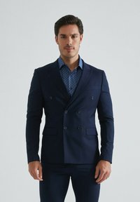 Blazer bleu marine à double boutonnage en tissu lisse, avec deux poches avant et six boutons. En dessous, une chemise à pois bleus apporte un contraste.