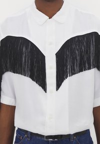 Camicia bianca a maniche corte con motivo a frange nere sul petto, chiusura con bottoni e colletto classico.