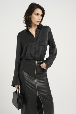 Camicia nera in satin con bottoni e due tasche sul petto, abbinata a una gonna nera in pelle con zip e una borsa nera con texture.