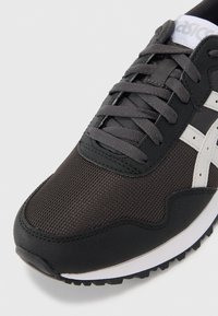 Baskets en mesh noires avec des accents gris, dotées d'une semelle en caoutchouc, d'un dessus texturé et d'un système de laçage traditionnel. Les détails incluent un logo blanc.