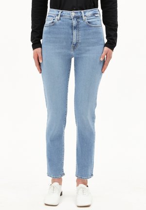 Helle blaue High-Waist-Jeans mit tailliertem Schnitt, ausgestattet mit einem Fünf-Taschen-Design und subtilen Ausbleichungen. Kombiniert mit weißen Schuhen.