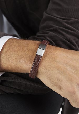 Handgelenk mit einem braunen geflochtenen Lederarmband und einem silbernen Magnetverschluss, das auf einer dunklen, strukturierten Stoffoberfläche liegt.