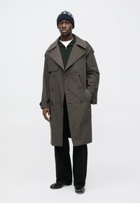 Gray double-breasted trench coat with large notched lapels, button accents, and side pockets, paired with a navy knitted hat and black pants.