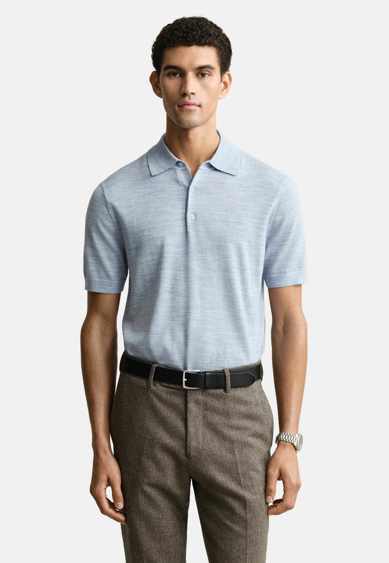 Homme portant un polo léger bleu clair à manches courtes, rentré dans un pantalon marron à texture avec une ceinture noire et une montre en argent, se tenant contre un fond blanc.