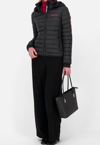 Sort dunjakke med lynlås foran og hætte, båret over en rød top. Sorte bukser med vide ben og en mønstret sort håndtaske fuldender outfittet.