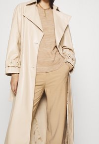 Beige, glänsande trenchcoat med stora slag och knäppta ärmmuddar, i kombination med en stickad beige tröja och krämfärgade byxor.
