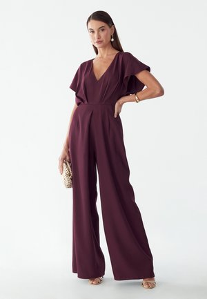 Donna in piedi che indossa una tuta prugna a gamba larga con maniche svolazzanti, tiene una pochette texturizzata e indossa orecchini e braccialetti dorati.