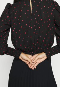 Blouse noire à motifs de cœurs rouges, poignets froncés, manches longues bouffantes, mains croisées dans le dos.