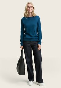 Blauwe gebreide trui, donkere spijkerbroek en witte sneakers. Houdt een zwarte tote bag vast. Eenvoudige ronde hals, lange mouwen, losse pasvorm.