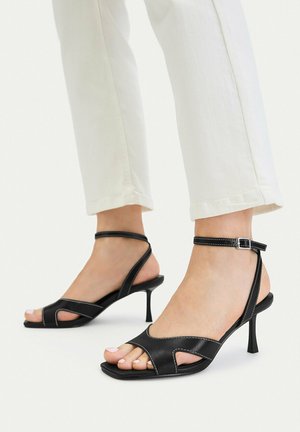 Füße in schwarzen, geschnürten Sandaletten mit Absatz und Knöfelriemen, kombiniert mit weißen, gekürzten Hosen vor hellem Hintergrund.