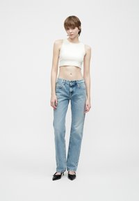 Persoană tânără îmbrăcată în blugi albaștri, top scurt fără mâneci alb și tocuri ascuțite negre, stând în fața unui fundal alb simplu.