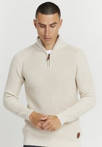 Beige strikkegenser med høy krage og halv glidelås, med ribbestrikkede mansjetter og kant, samt et lite lærlogo-merke på undersiden.