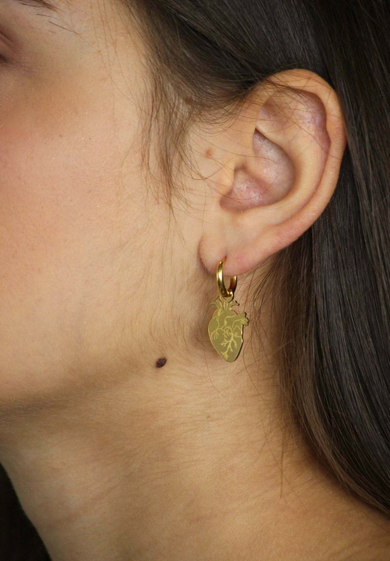 Pendientes de aro de oro con un colgante en forma de corazón con detalles intrincados, colgando del lóbulo de la oreja sobre un tono de piel neutro.