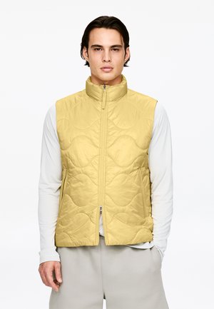 Gesteppte gelbe Weste mit hohem Kragen und Frontreißverschluss, getragen über einem hellgrauen Langarmshirt. Glatte Textur, lässiges Design.