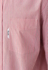 Chemise rayée rouge et blanche, avec une poche poitrine gauche et une étiquette cousue. Fabriquée en tissu léger à texture lisse.