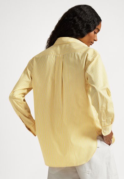 Sieviete ar gariem cirtainiem matiem, valkājot dzeltenu un baltu vertikālu svītru garroku kreklu un baltas bikses, skatoties no muguras.