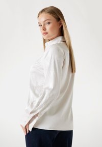Blouse blanche à manches longues en satin avec un col. Présente des broderies décoratives en argent sur les manches et une coupe décontractée.
