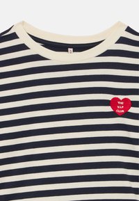 Tamsiai mėlynas ir kreminės spalvos dryžuotas marškinėlių modelis su ribuota apvaliomis kaklo apykaklė. Yra raudonas siuvinėtas širdies lipdukas su baltu užrašu „THE V.I.P. CLUB“.