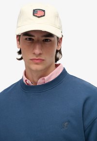Gorra de béisbol crema con parche negro que presenta una bandera estadounidense y el texto "SUPERDRY". El modelo lleva un sudadera azul y una camisa de color rosa debajo.