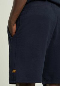 I pantaloni corti in cotone blu navy presentano un ampio elastico in vita, tasche laterali e una piccola etichetta con il logo vicino all'orlo, mostrando una consistenza liscia e uniforme.