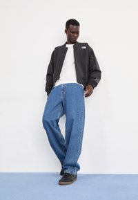 Giacca bomber nera, camicia bianca, jeans in denim blu larghi. Il modello indossa scarpe nere. La giacca presenta un logo sul petto e dettagli con zip.