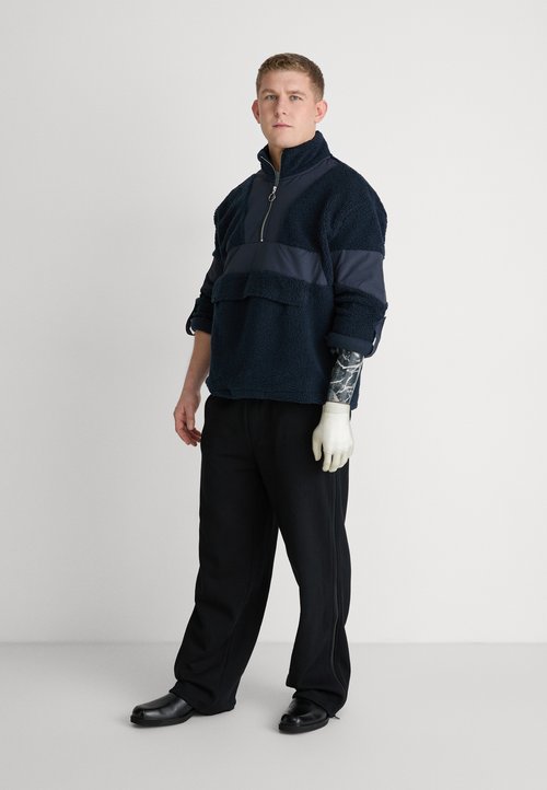 Námornícky fleecový pulover s polovičným zipsom a spodnou prednou kapsou, skombinovaný s čiernymi širokými nohavicami a čiernymi topánkami. Na rukách sú svetlé rukavice.