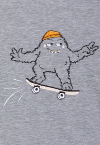 Un tissu gris arbore un monstre de dessin animé brodé sur un skateboard, portant un chapeau orange, avec une expression ludique et des détails texturés.
