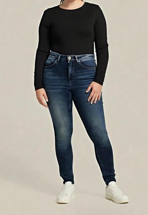 Person, die dunkelblaue, hoch taillierte Skinny-Jeans, ein schwarzes Langarmshirt und weiße Turnschuhe trägt, steht vor einem einfarbigen Hintergrund.