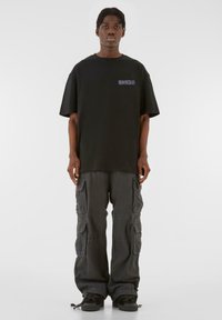 Tricou negru supradimensionat cu un grafic pe piept, asortat cu pantaloni cargo largi de culoare închisă, care au numeroase buzunare și tivuri cu șnur.