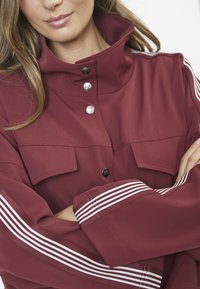 Burgundy takki, jossa on korkea kaulus, hopeiset napit, kaksi rintataskua ja valkoiset raidat hihoissa; sileä pinta.