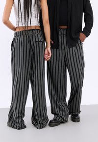 Pantaloni largi cu dungi negre și gri, cu bandă elastică la talie, având detalii cu buzunare. Materialul pare neted și ușor.
