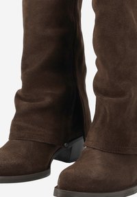 Stivali alti in suede marrone, con un leggero tacco, caratterizzati da una parte superiore piegata, una texture sottile e una chiusura laterale con un piccolo dettaglio a bottone.
