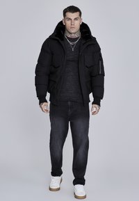 Black puffer jacket with a fur-lined hood, paired with a dark gray textured shirt and black jeans. White sneakers with a brown sole.