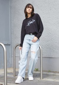 Czarna koszula z długim rękawem z logo „Levi's”, jasnoniebieskie podarte dżinsy i białe trampki. Stoi obok metalowej poręczy na chodniku.