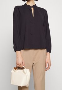 Zwarte blouse met een hoge kraag en een opensnijding, gecombineerd met beige broeken. Houdt een witte handtas vast met een zachte structuur en een houten handvat.