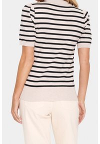 Beige kortärmad topp med svarta horisontella ränder; ribbad krage och fåll; mjukt stickad textur. Modellen har på sig ljusfärgade byxor.