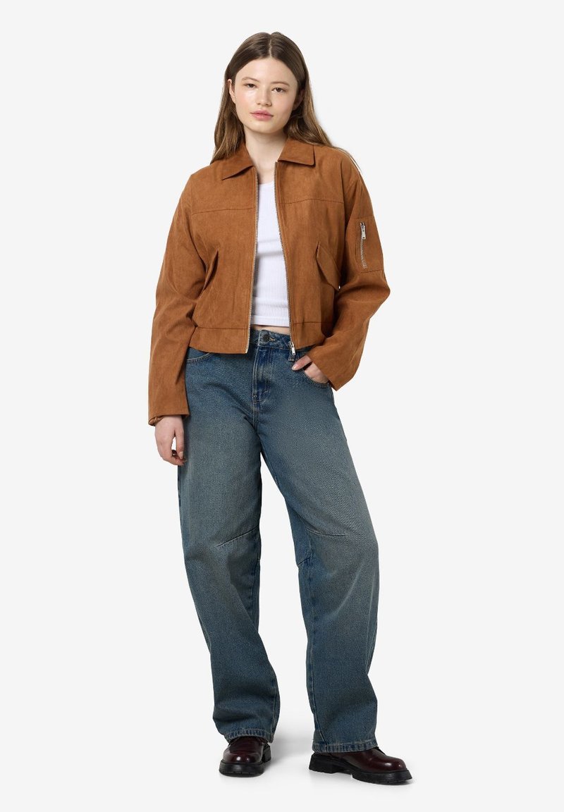 Veste en cuir suédé brune courte avec un col, poche zippée sur la manche, associée à un jean en denim bleu à coupe décontractée et des bottes noires.