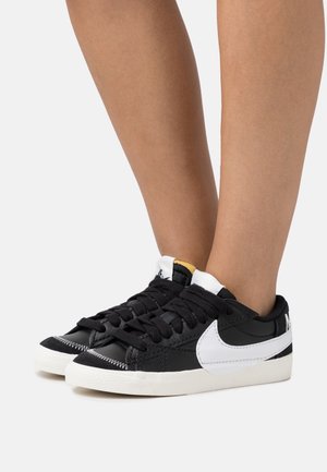 Jambes portant des baskets basses noires avec logos swoosh blancs, semelles épaisses blanches et lacets noirs, debout sur une surface blanche.