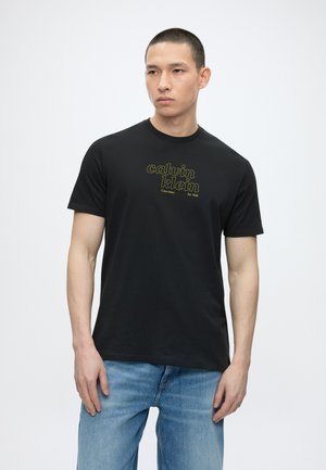 Άντρας που φοράει μαύρο t-shirt Calvin Klein με κίτρινο λογότυπο και μπλε τζιν, στέκεται μπροστά σε απλό λευκό φόντο.