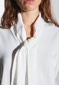 Blusa bianca con dettagli al collo legati, tessuto drappeggiato, texture liscia e vestibilità rilassata, che enfatizzano un design minimalista.