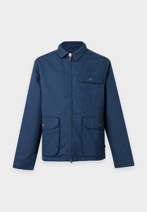 Homme portant une veste bleue, une chemise vert foncé, un pantalon beige clair et des chaussures marron, tenant un sac fourre-tout beige avec des sangles marron.