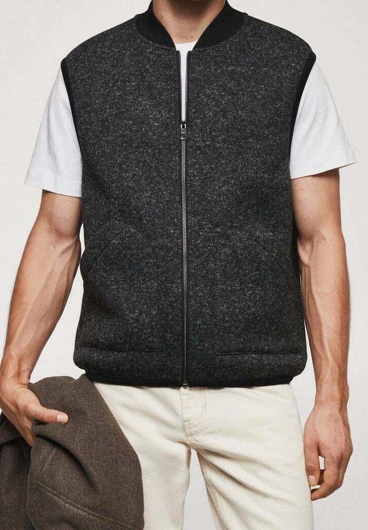 Homme portant un gilet sans manches zippé gris foncé par-dessus une chemise blanche à manches courtes et un pantalon clair, tenant une veste marron pliée dans la main gauche.
