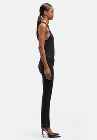 Szczupła kobieta z długimi ciemnymi włosami związanymi w kucyk, ubrana w czarny bezrękawnik, czarne dopasowane spodnie i czarne szpilki, stojąca bokiem.