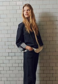 Giacca di jeans in denim scuro con polsini ripiegati e strisce multicolori. Abbinata a jeans scuri dal fit ampio e una camicia chiara.