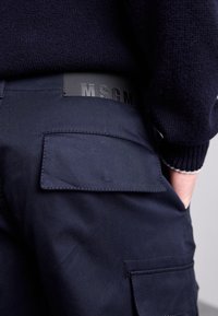 Mørkeblå bukser med en baglomme og et sort logomærke ved taljen, lavet af glat stof med et struktureret design.