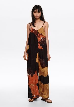 Kobieta ubrana w bezrękawnikowy kombinezon z szerokimi nogawkami w abstrakcyjny wzór w kolorach czarnym, pomarańczowym i musztardowym, stojąca boso na tle jednolitej białej ściany.