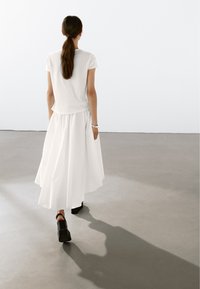 Hvid t-shirt parret med en lang, flydende hvid nederdel. Modellen bærer sorte blokhælede sko. Glatte teksturer; minimalistisk design.