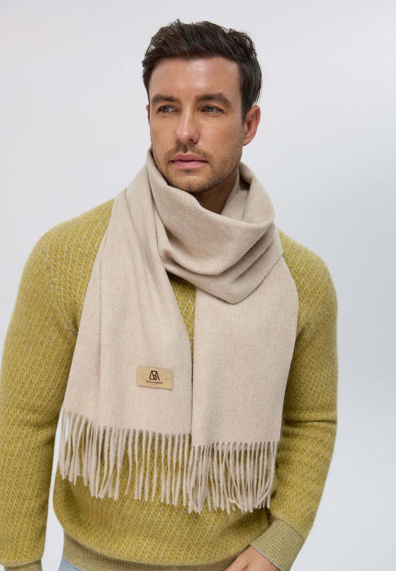 Sciarpa beige con dettagli in frangia, realizzata in un tessuto morbido. Indossata in modo morbido sopra un maglione verde oliva dalla trama, con un colletto rotondo.