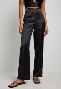 Pantalons larges en satin noir avec une texture lisse, dotés d'une taille haute et de poches latérales, associés à des tongs noires.