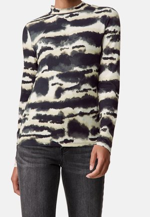 Kvinde iført en sort og hvid langærmet top med abstrakt mønster, kombineret med mørkegrå jeans, stående mod en ensfarvet baggrund.